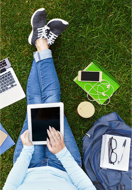 Female student sitting in the grass with computer, phone, and tablet
